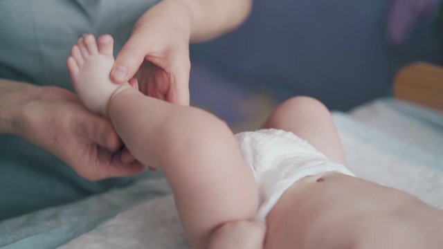 small baby boy in white diaper lies on changing table and professional nurse performs legs massage close view