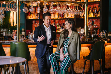 Portrait of two professionally and well-dressed young Asian individuals out on a date at a classy bar together. One is a Chinese man in a suit with his elegant Indian woman date -- they are talking.