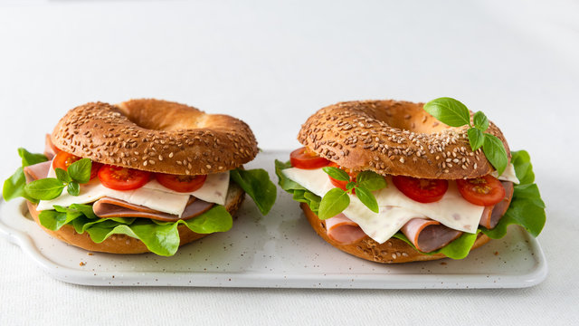 Bagel sandwich with ham, cheese, salad and cherry tomatoes on white chopping board on white background.