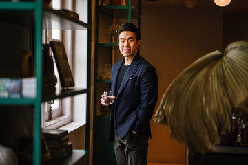 Portrait of a handsome, good looking, young Chinese Asian man in a casual and well-tailored suit, tee shirt as he stands by the window of a well-decorated room while holding a glass during the day.