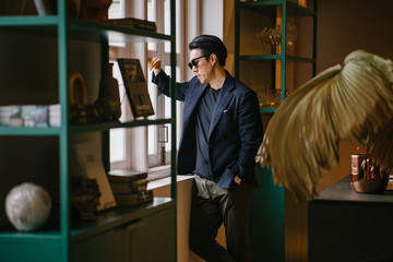 Fototapeta na wymiar Portrait of a handsome, good looking young Chinese Asian man in a casual and well-tailored suit, tee shirt and sunglasses as he gazes out the window of a well-decorated room during the day.