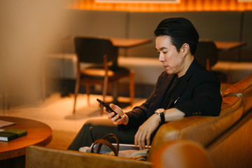 Fototapeta na wymiar Portrait of a handsome, confident and well-dressed young Chinese Asian man checking his smartphone as he sits on a leather sofa in a well-appointed sofa during the day.