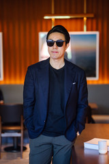 Portrait of a handsome, young and fit Chinese Asian man in a casual suit and sunglasses. He is standing and smiling in a warm, well-decorated room during the day. He is neat, dashing and confident.