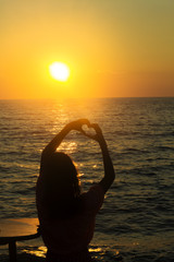 silhouette of  girl on a sunset background that holds heart shaped fingers by the sea