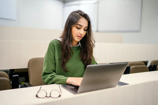 Portrait of a beautiful, young and intelligent-looking Indian Asian woman student wearing a white shirt and green tracker smiling as she works on her laptop in a university classroom.