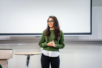 Portrait of a young, beautiful, attractive and intelligent-looking Indian Asian woman wearing spectacles in a sweater giving a business presentation to an audience. - Powered by Adobe