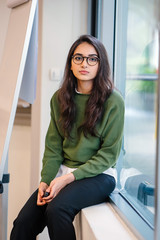 Portrait of a young, educated, confident and intelligent-looking Indian Asian teacher woman smiling as she sits in a seminar room by the window during the day in a preppy outfit.