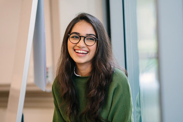 A close up head shot portrait of a preppy, young, beautiful, confident and attractive Indian Asian woman in a green sweater and spectacles in a classroom or office. She is smiling happily. - obrazy, fototapety, plakaty