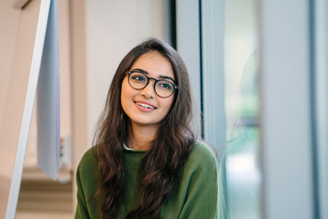 A close up head shot portrait of a preppy, young, beautiful, confident and attractive Indian Asian woman in a green sweater and spectacles in a classroom or office. She is smiling happily.