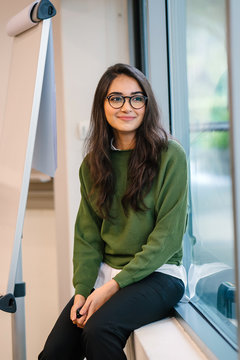 Portrait of a young, educated, confident and intelligent-looking Indian Asian student girl smiling as she sits in a seminar room by the window during the day in a preppy outfit.