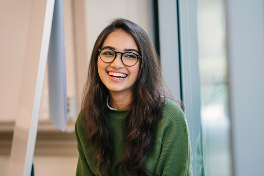 A close up head shot portrait of a preppy, young, beautiful, confident and attractive Indian Asian woman in a green sweater and spectacles in a classroom or office. She is smiling happily.