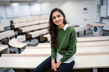 Portrait of a beautiful, young and attractive Indian Asian business woman smiling as she sits in a seminar room during the day. She is wearing a preppy green sweater over a white shirt and jeans. - obrazy, fototapety, plakaty