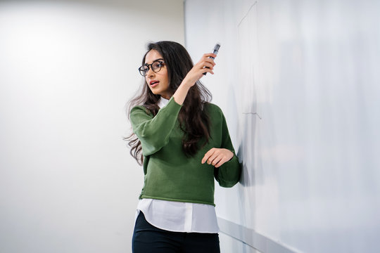 Portrait Of A Young, Beautiful, Attractive And Intelligent Looking Indian Asian Businesswoman Explaining A Concept By Sketching On A Whiteboard. She Is Wearing A Preppy Green Sweater And Glasses.