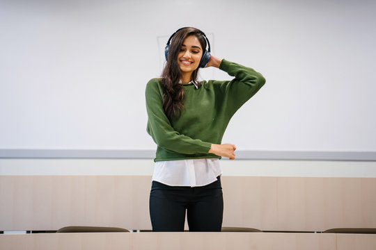 Portrait of a beautiful, young and attractive Indian Asian woman student in a preppy outfit smiling as she dances to music that she is streaming on her phone to her wireless headset in a classroom.