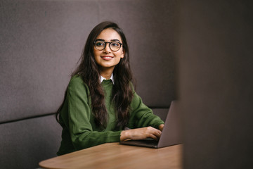 Portrait of a young, confident, intelligent-looking and attractive Indian Asian graduate student. She is smiling as she checks her smartphone. She is sitting in a study booth on her campus.
