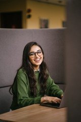 Fototapeta na wymiar Portrait of a young, confident and attractive Indian Asian student working or studying on her laptop as she sits in a booth. She is dressed in a preppy sweater and shirt and glasses.