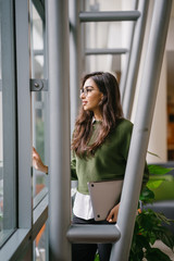 Portrait of a beautiful, young, elegant and attractive Indian Asian woman student in a preppy green sweater and spectacles. She is relaxing in a glass building during the day.
