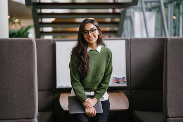Portrait of a young, beautiful, intelligent and attractive Indian Asian MBA student smiling as she leans in a discussion booth in her campus. She is wearing a preppy outfit and smiling confidently. - obrazy, fototapety, plakaty