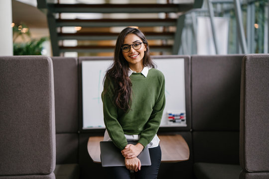 Portrait of a young, beautiful, intelligent and attractive Indian Asian MBA student smiling as she leans in a discussion booth in her campus. She is wearing a preppy outfit and smiling confidently.