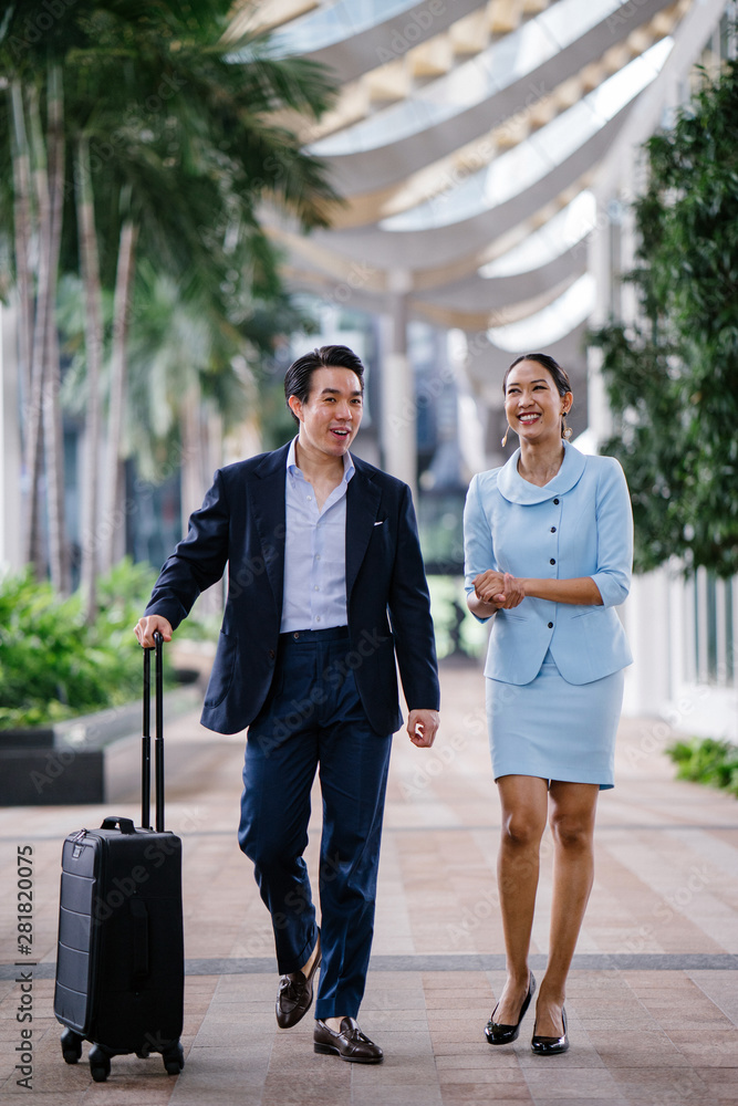 Wall mural Portrait of two well-dressed and professional Asian business people walking together in a modern city during the day. A Chinese man in a dark navy suit is talking to his female companion as they walk.