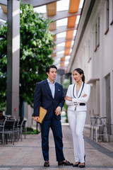 Fototapeta premium Portrait of two young, confident and attractive Asian business people standing together in a modern city during the day. They are both professionally dressed in suits and are smiling.