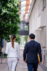 Two young professionals walk side by side. One is a Chinese Asian man in a suit and the other a woman in a white suit -- they are both engaged in conversation as they walk in the city together.