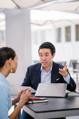 A Chinese Asian manager in a suit has a meeting with his colleague, a woman in a pale blue suit. He is conducting a performance appraisal during this meeting. They are smiling and talking.