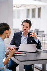 A Chinese Asian manager in a suit has a meeting with his colleague, a woman in a pale blue suit. He...
