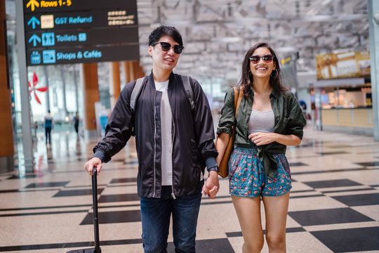 A young and photogenic Asian couple (Korean man, Indian girlfriend in sunglasses) smile as they walk in an airport. They are pulling their luggage behind them on their way for a vacation overseas.