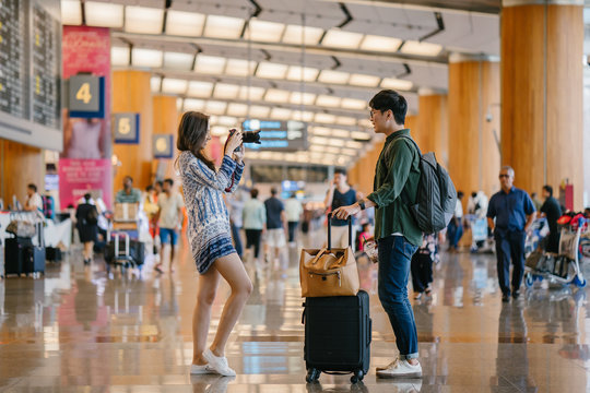 Portrait of a young, diverse and casually dressed interracial Asian couple (a Korean man and Indian woman) standing and taking pictures of one another in a futuristic airport during the day.