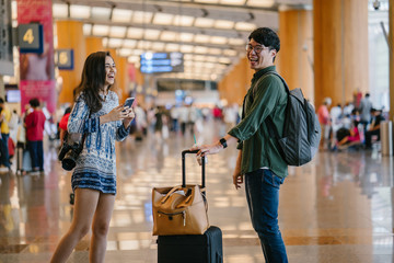 A young interracial diverse couple casually dressed as they stand in the middle of an airport during the day. A young,handsome Korean man and his Indian woman companion are laughing out loud together.
