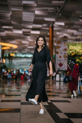 Full body portrait of a young and attractive Indian Asian woman twirling in her casual and comfortable black dress and white sneakers. She is in a futuristic and modern airport or mall.