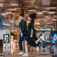 A diverse Asian pair (Indian woman and a Korean man) smile and laugh as they leap and jump together in sync in a modern airport during the day. They are casually dressed and are having great fun.