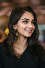 A close up portrait of a beautiful, young, attractive and photogenic Indian Asian girl during the day in the city. She is wearing a casual black dress and smiling radiantly.