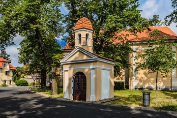 Fototapeta na wymiar Panensky Tynec, Czech Republic - July 15 2019: Chapel of John of Nepomuk with yellow and white facade standing in the street. Green grass, trees around. Sunny summer day with blue sky and clouds.