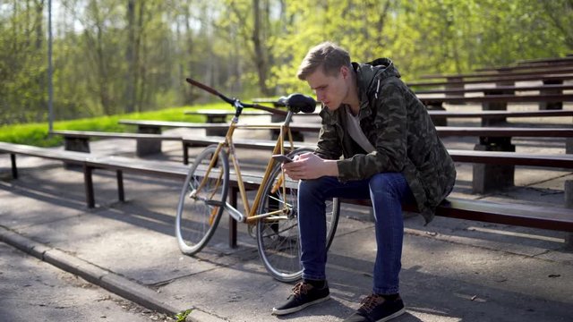 A young man sitting on a bench in a bleacher next to a yellow bicycle and checking his cell phone