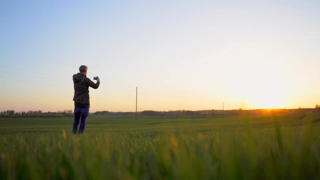 View of a young man taking photos with his cell phone in a field at sunset with yellow and golden tones in the sky