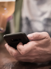 close up of man hand hold smartphone, drinking beer and reading message at bar or pub