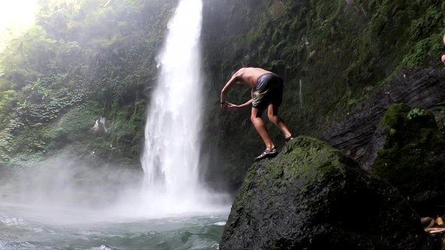 Slow Motion Shot of a man doing a front flip off of a rock and into the swimming hole at the base of the gushing NungNung Waterfall in Bali, Indonesia.