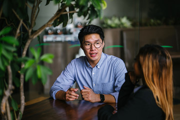 A young Asian business woman during a consultation with a young Korean Asian man for business advice. They are sitting in a trendy coworking space during the day and having a business discussion.