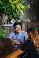 A confident and handsome young man is interviewing a female candidate for a job. They are sitting together in a table at a trendy coworking space. He is smiling and having a relaxed conversation.