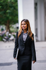 Fototapeta premium Portrait of a beautiful, young and attractive Singaporean Malay Asian woman in a business suit walking in a city in Asia during the day with her laptop computer. She is smiling confidently.