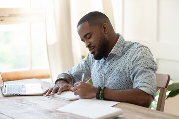 Focused african man student freelancer making notes studying with laptop