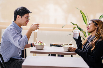 A pair of diverse Asian colleagues (Malay woman, Korean man) smile and toast one another to celebrate an achievement or promotion over lunch during the day. They are both smiling happily.
