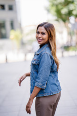Portrait of a young, attractive, beautiful, tanned and sexy Malaysian Asian woman in a denim jacket and a casual outfit strolling and walking in a street in the city during the day. She is smiling.