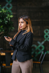 Fototapeta premium A young, tanned, attractive and confident Southeast Asian business woman in a suit smiles as she checks and messages on her smartphone. She is standing in a trendy coworking space or cafe in the day.
