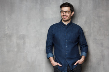 Inspired millennial man wearing trendy glasses, looking away while standing against gray wall