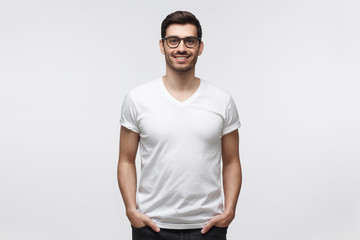 Handsome smiling man in white t-shirt and trendy eyeglasses, standing with hands in pockets, ...