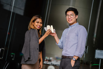 Business portrait of two diverse Asian business people (colleague meeting for a snack) standing by the cafe. One is a Korean man,the other a Malay woman. They're both smiling and holding their treats.