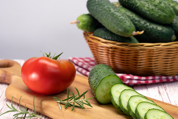 On a wooden board is a red tomato and a sliced cucumber. Near a basket with fresh cucumbers. Summer vegetables, eco products.